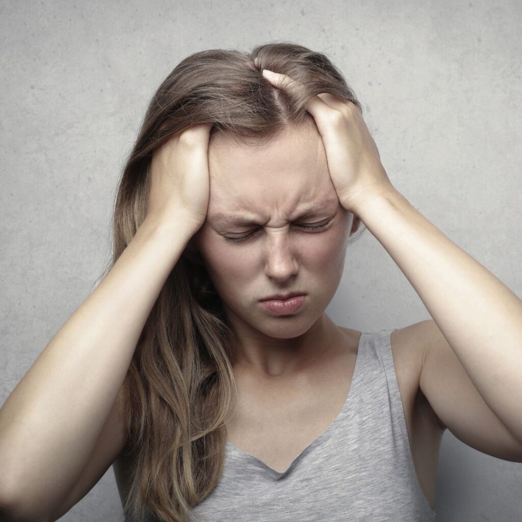 Young woman experiencing a severe headache or migraine, pressing her temples with both hands and eyes closed in pain, standing against a neutral gray background.
