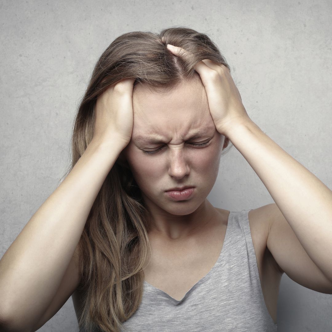 Young woman experiencing a severe headache or migraine, pressing her temples with both hands and eyes closed in pain, standing against a neutral gray background.